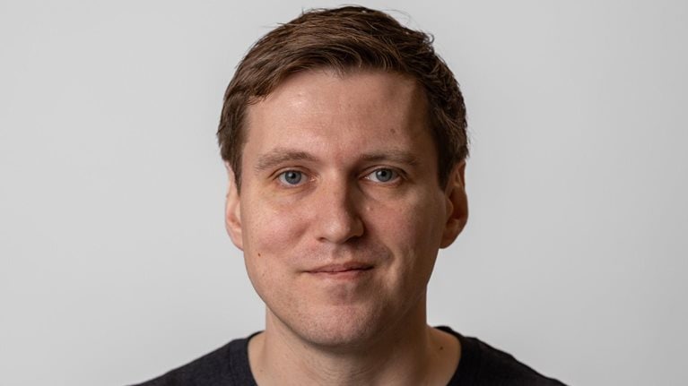 Philip Johnston with short brown hair and blue eyes looks directly at the camera with a slight, relaxed smile. He wears a dark shirt and is set against a plain, light-colored background.