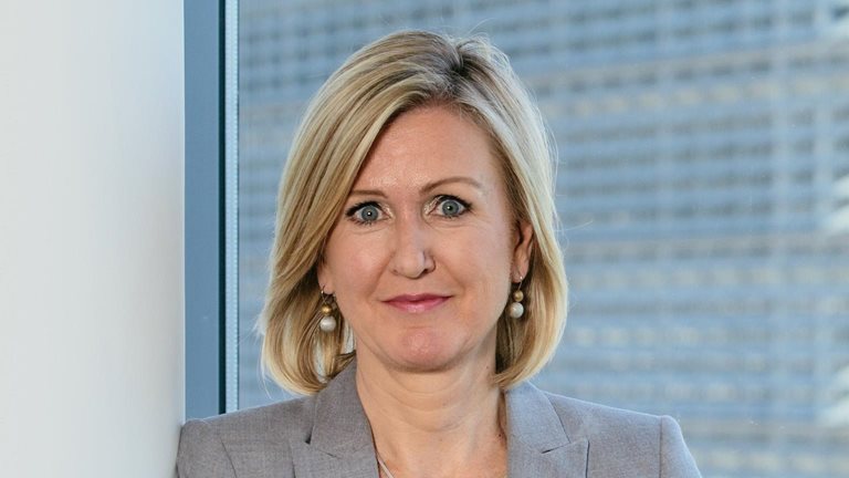 Portrait of Ann Mettler leaning against a pillar with large windows behind her in a modern office building.