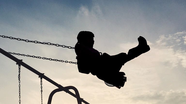 A silhouetted child soars through the air on a swing set. The setting sun casts a warm glow on the scene, creating a quiet and nostalgic atmosphere.