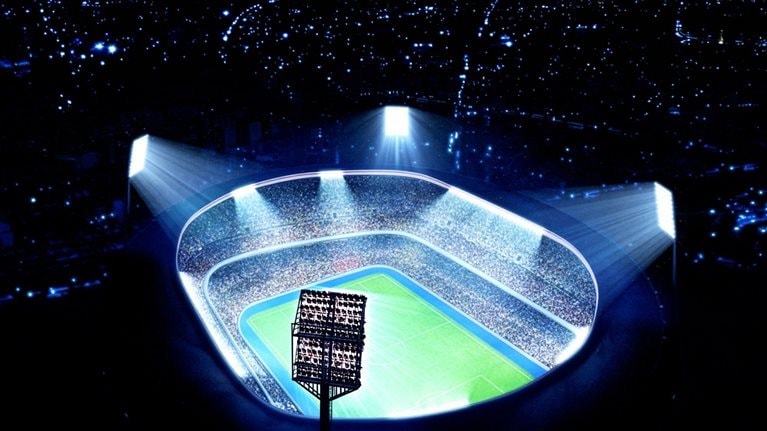 A brightly lit, modern soccer stadium packed with spectators is shown from an aerial perspective, with powerful floodlights illuminating the field at night. The scene emphasizes scale and energy, highlighting a vibrant sports arena surrounded by darkness.