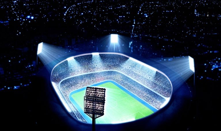 A brightly lit, modern soccer stadium packed with spectators is shown from an aerial perspective, with powerful floodlights illuminating the field at night. The scene emphasizes scale and energy, highlighting a vibrant sports arena surrounded by darkness.
