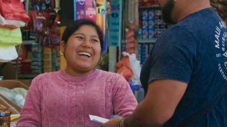 Photograph of a small grocery store checkout area showing a cashier wearing a pink sweater and gloves interacting with a customer in a blue shirt and cap. Shelves behind the cashier are stocked with various packaged food items, including biscuits, cereals, and snacks, highlighting a typical local market setting.