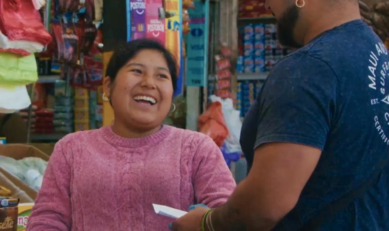 Photograph of a small grocery store checkout area showing a cashier wearing a pink sweater and gloves interacting with a customer in a blue shirt and cap. Shelves behind the cashier are stocked with various packaged food items, including biscuits, cereals, and snacks, highlighting a typical local market setting.