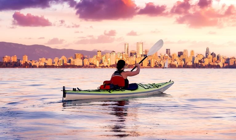 As the sun sets behind the Vancouver skyline, a woman paddles her kayak on a vast body of water. The vibrant sunset sky paints the horizon with shades of pink and purple, casting a mirror-like reflection on the water.