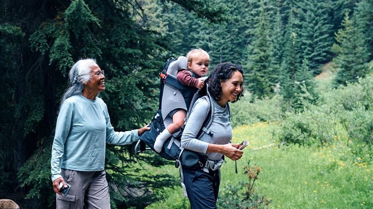 A smiling family, composed of a grandmother, grandfather, mother with a baby in a carrier on her back, and a young girl, treks through a forest, crossing a wooden bridge over a small stream. The grandfather assists the girl as they walk.