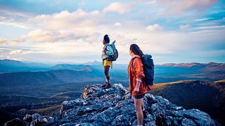 Two hikers stand on a rocky mountain peak, wearing backpacks and looking out over a vast, scenic landscape. The expansive view and soft light convey a sense of adventure, achievement, and connection with nature.