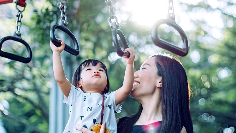A mother smiles as she holds her young child up while the child grasps a monkey bar in an outdoor playground.
