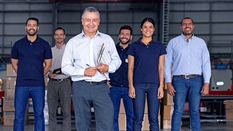 A man inside a warehouse leading a group of Latin American warehouse employees; he is holding a clipboard and the team is scattered behind him but all are smiling and looking at the camera